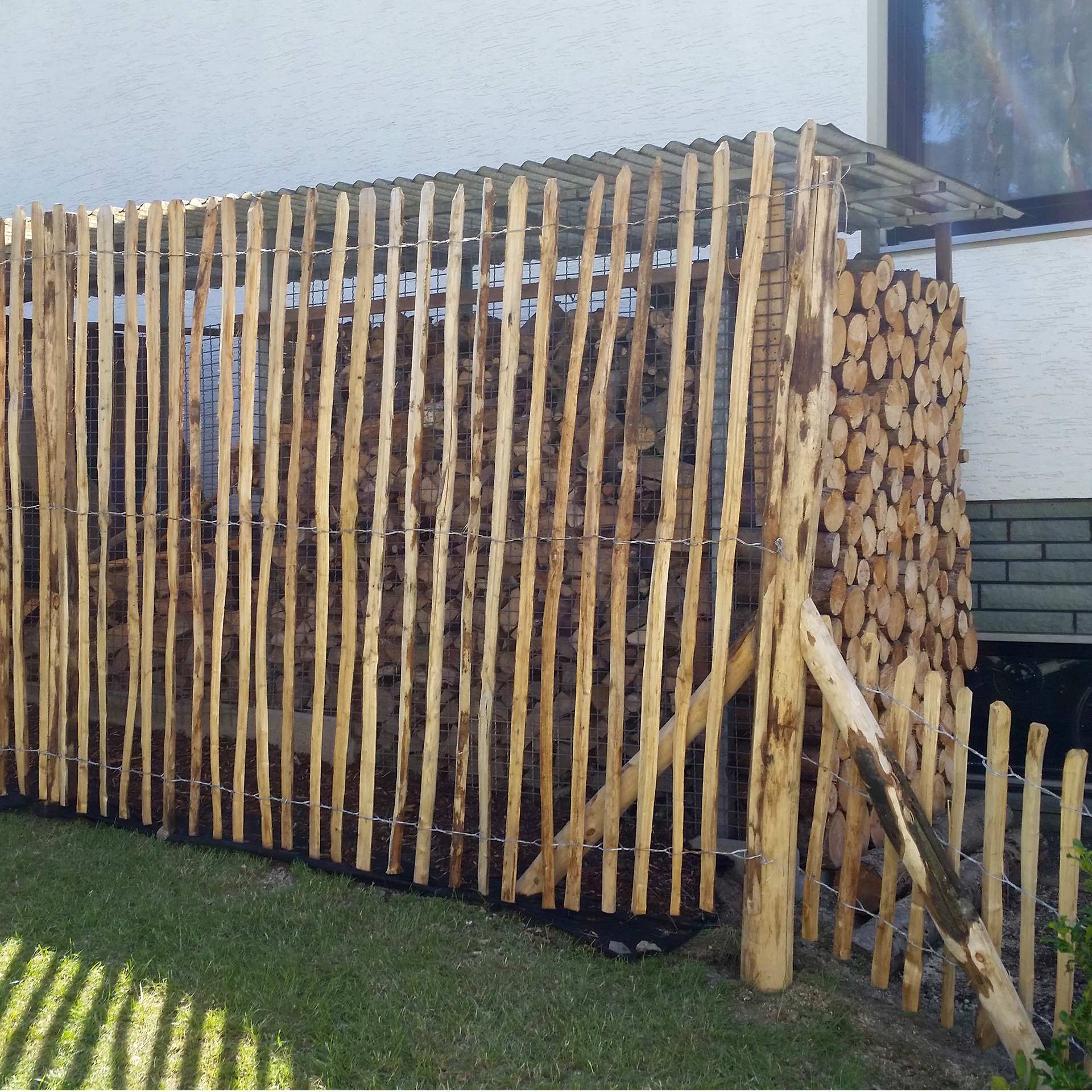 Staketenzaun als Sichtschutz vor Holzstapel an hauswand, Holzstapel, Holzstapel-Zaun, Sägewerkholz, Staketenzaun, Holzpfosten